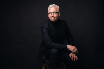 Serious businessman wearing glasses with white hair at the camera with a focused expression against a black studio background