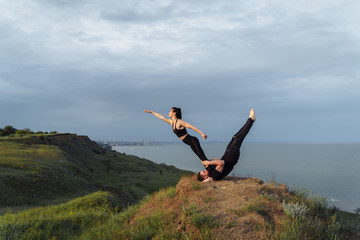 Sportive, strong athletes making acrobatic performances on the hill against sea during sunset. The athlete lying on his back and standing on the shoulder blades, holding by 
abdominal press