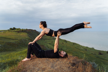 Young couple guy and girl doing acro yoga on the background of the sea. Man lying on the grass and balancing woman on his legs. Dressed in black sportswear. Woman is flying in the air