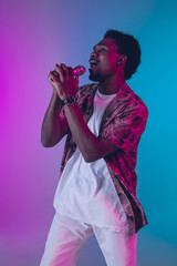Fototapeta na wymiar African-american male singer portrait isolated on gradient studio background in neon light. Beautiful male model with microphone. Concept of human emotions, facial expression, ad, music, art.