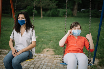 Girl and woman with face mask enjoying swinging in the park. Portrait of girl wearing protection mask while swinging at playground with adult woman.