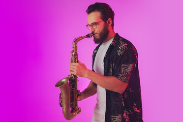 Fototapeta premium Young caucasian jazz musician playing the saxophone on gradient pink studio background in neon light. Concept of music, hobby, festival. Joyful, cheerful attractive guy. Colorful portrait of artist.