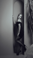 Black and white shot of sexy young girl wearing short off shoulder black dress and leaning on wall