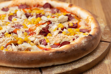 hearty italian pizza on a wooden board. crispy cheese pizza with hunting sausages, pepper, chicken and corn