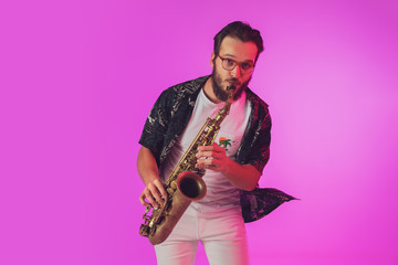 Naklejka premium Young caucasian jazz musician playing the saxophone on gradient pink studio background in neon light. Concept of music, hobby, festival. Joyful, cheerful attractive guy. Colorful portrait of artist.