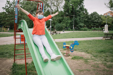 Happy girl with antivirus mask going down the slide with hands wide open. Girl enjoying the freedom of sliding at the playground after COVID-19 regulations are relaxed