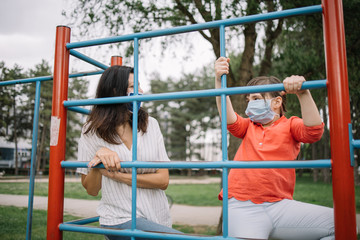 Nice girl with face mask climbing fence at play park while woman is assisting her. Lady and child behind climbing fence at playground protected against coronavirus