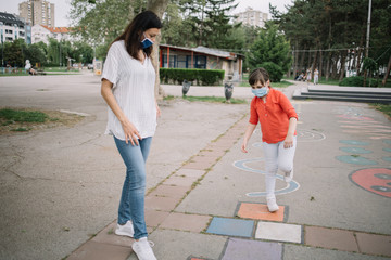 Brunette woman watching girl playing games on asphalt with masks on their faces. Lady and child...