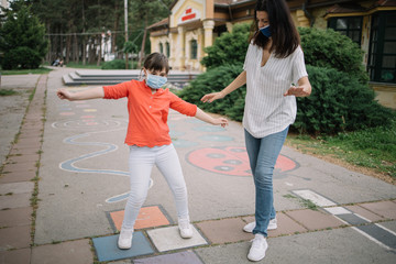 Young woman and cute girl at playground markings using protection masks. Lady showing child how to hold balance while playing games on asphalt.