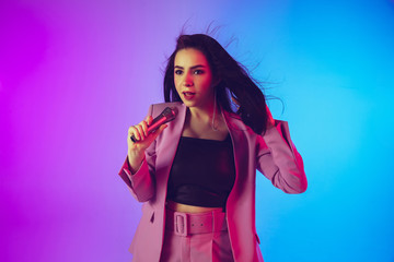Caucasian female singer portrait isolated on gradient studio background in neon light. Beautiful female model in pink wear with microphone. Concept of human emotions, facial expression, ad, music, art