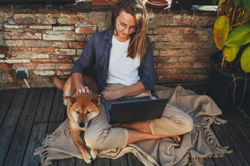 Young freelancer woman in casual clothes and eyeglasses sits at home sunny terrace with shiba inu dog relaxed with legs crossed using laptop computer watching video or working remotely