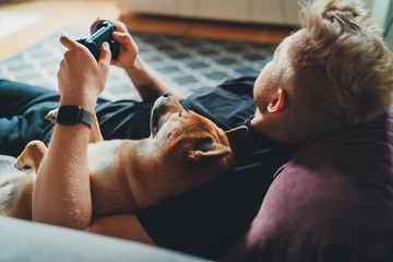 Young hipster guy playing video games at home holding game joystick relaxing on couch with his best friend dog, Home Leisure Concept