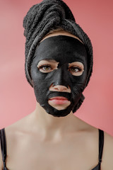 Young woman apply black cosmetic fabric facial mask on pink background. Face peeling mask with charcoal, spa beauty treatment, skincare, cosmetology. Close up