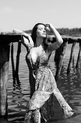 Dark-haired woman posing at the wooden pier at the lake. Woman in summer outfit with her back turned to the wooden footbridge of Lake Neusiedl. Sun reflections at the boathouse in the background