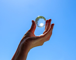 Female hand holding a transparent crystal ball with city and trees reflection. Round glass sphere with sunbeams against a blue sky background. Bright summer view.