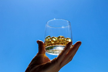 Hand holds a glass with fish oil pills. Transparent round golden capsules of vitamin on a blue sky background. Concept of treatment, healthy nutrition and prevention of health.