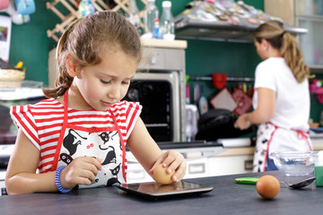 bambina con maglia a righe pesa un uovo nella bilancia elettronica mentre la madre cucina sullo sfondo 