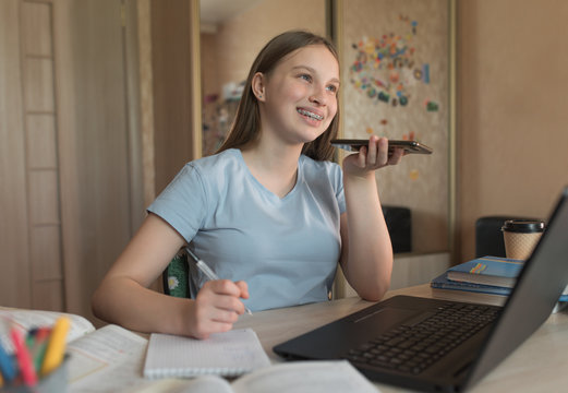 Happy smiling, teenager girl talking and recording a voice message, smartphone, laptop lessons at home in hand. E-education and distance learning through the Internet and video communications.
