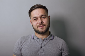 Young Bearded Dark Haired Man In Gray Stylish T-Shirt On Background, Portrait Of A Serious Man Who Looks Straight At The Camera. Close Up