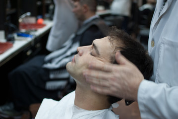 Barber finishing young man haircut and gives face massage at a barbershop