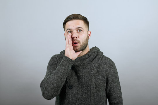 Young Bearded Dark Haired Man In Black Stylish Shirt On Gray Background, Talking Male Holds Hand To His Mouth And Speaks. The Concept Of Chatter, Gossip, Talk