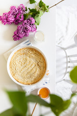 Tasty cheesecake in a white dish on a table with lilacs, top view, flat lay