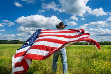 Man wearing cowboy hat standing in grassy agricultural field with American flag blowing in the...