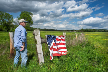 Farmer standing in grass agricultural field, American flag draped over barbwire fence, blue sky...