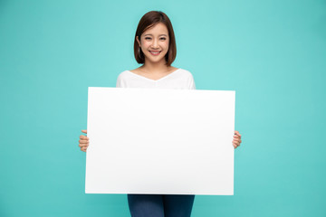 Smiling happy Asian woman holding and standing behind big white poster isolated on light green...