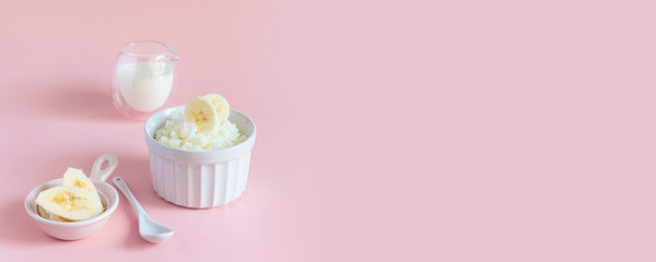 Tasty rice milk porridge with banana in white mask and milk in milkman on pink background with copyspace