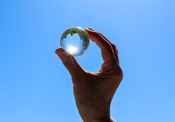 Female hand holding a transparent crystal ball with city and trees reflection. Round glass sphere with sunbeams against a blue sky background. Bright summer view.