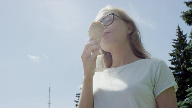 Pretty becpectacled blonde girl eats ice-cream cone thoughtfully looking away from camera in front of blue clear sky. Low-angle shot.