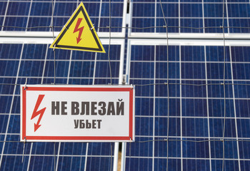 Solar power farm station on a background selective focus on a yellow triangular high voltage warning sign. The inscription in Russian Do not come near kill 