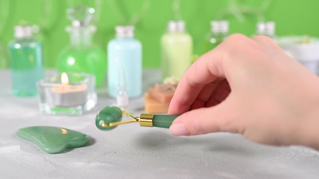 lady hand with manicure takes green jade roller from white surface table against blurry containers and spa equipment closeup
