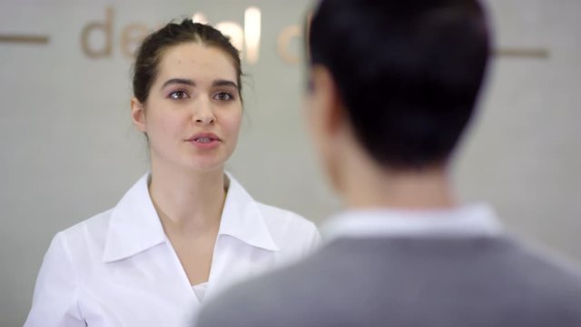Medium shot of cheerful young female medical receptionist greeting patient and giving her registration form