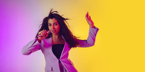 Caucasian female singer portrait isolated on gradient studio background in neon light. Beautiful female model in pink wear with microphone. Concept of human emotions, facial expression, ad, music, art