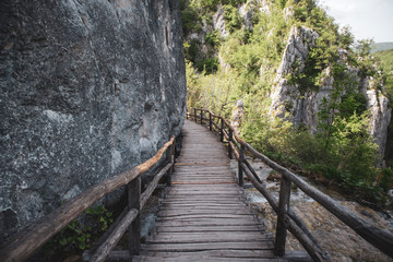 Fototapeta na wymiar Old, wooden bridge crossing over Plitvice lakes national park waterfalls, beneath massive stone rock