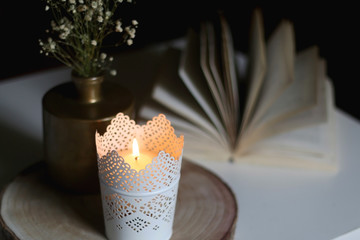 Candle holder with lit candle, golden vase with gypsophila flowers and open book on a table. Selective focus, moody lighting.