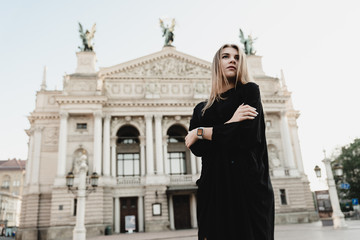 Fototapeta premium A beautiful young woman in a black dress with blond hair walks around the city with a bag with red flowers, filtered image, flare sunshine