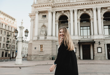 Fototapeta premium A beautiful young woman in a black dress with blond hair walks around the city with a bag with red flowers, filtered image, flare sunshine