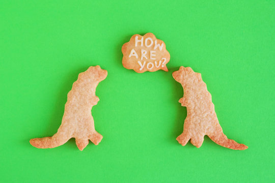 Homemade Shortbread Cookies In Shapes Of Dinosaurs With Callout Cloud With Text ‘How Are You?’ On Green Background, Top View.