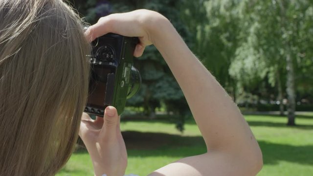 Young blonde woman with camera trying to take pictures of green lawn and birch trees in park on sunny day. Close shot from behind.