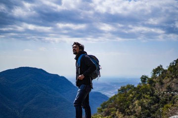 Young handsome indian boy standing on the top of the mountain with a bag pack while looking upwards in motivation