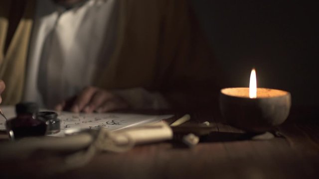 A medieval man uses an old pen with ink to write on an a paper lit by a candle,slow motion