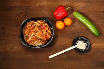 Black plate with funchose and chicken with vegetables under pad Thai sauce on a wooden table and Kujut seeds and chopsticks.