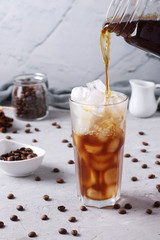 Pouring cold coffee into tall glass with ice cubes on light grey background, Refreshment drink, Vertical format, Closeup
