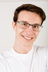 Fototapeta premium Young smiley guy with dark hair, glasses and a T-shirt making selfies on a white wall.