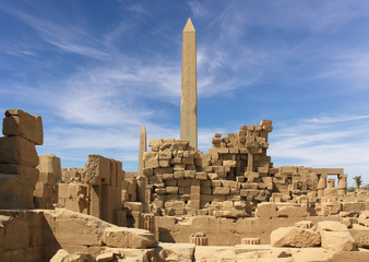 An obelisk and many stone columns in the ancient temple of Karnak in the Egyptian city of Luxor. In the foreground are many stones and walls. There are beautiful clouds in the blue sky.