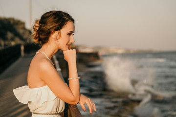 Beautiful young smiling girl in the white dress in greek style walking along the sea promenade. Summer, sunset time. Bride wedding at sea 