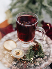 Hot Christmas mulled wine. The glass is on an iron plate, and there is a compass next to it. Red textiles. Forest, winter. Fir branches. Garland. Spices, Banyan, cinnamon. The table is wood.